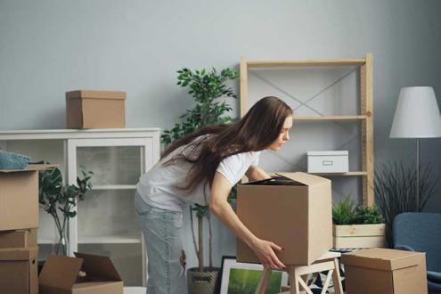A woman unpacking cardboard boxes in a new home, surrounded by plants, furniture, and home decor items